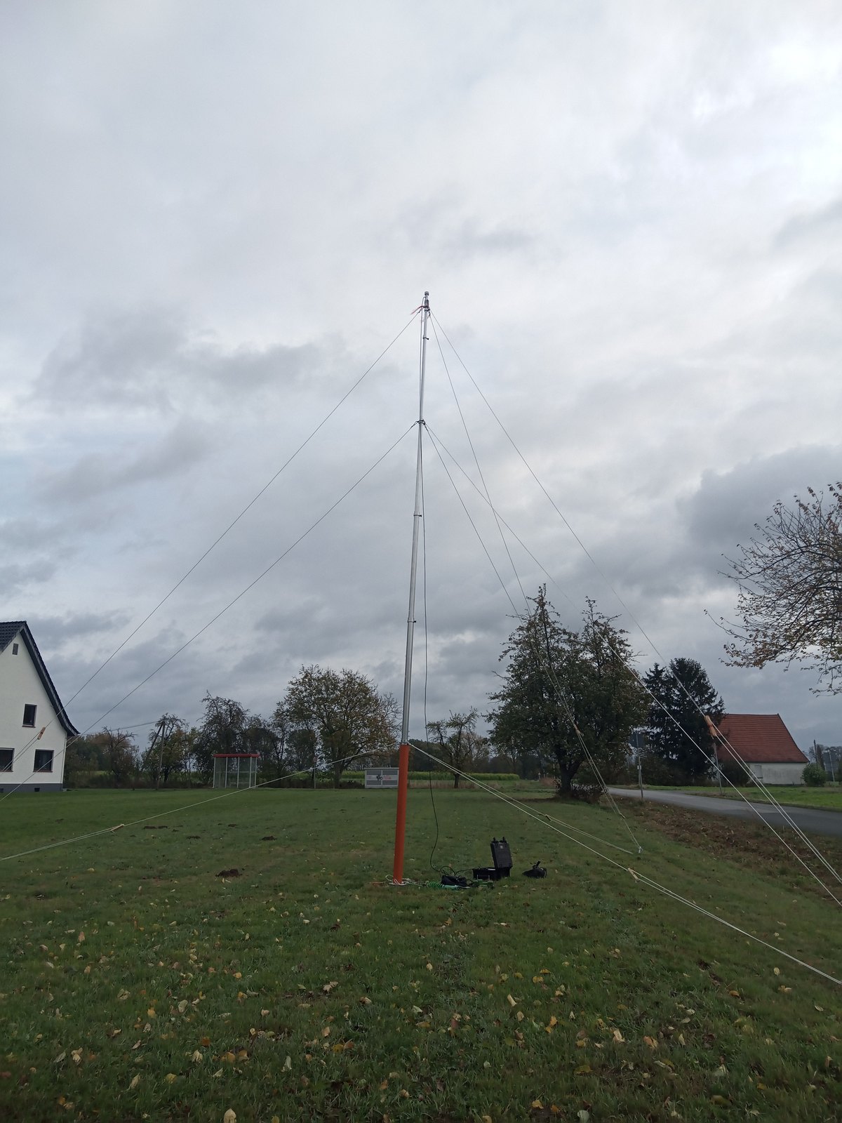 Mobiler Windmessmast einer Windreaktionsmessung auf einer Wiese an einem trüben Oktobertag, im Hintergrund Laubbäume, die noch weitgehend belaubt sind